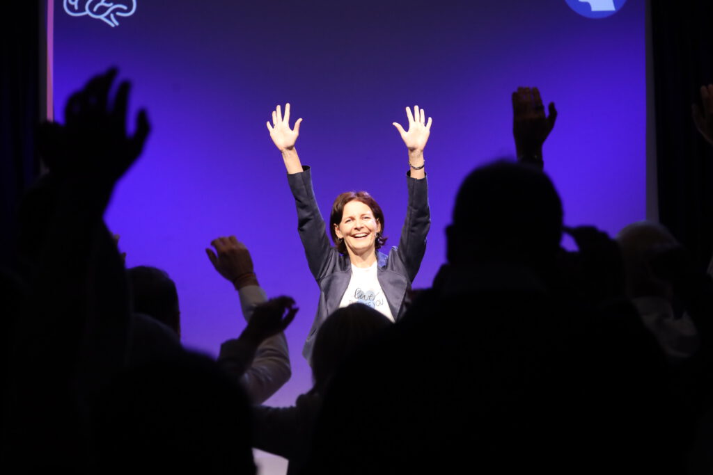 Kirsten Wunderle auf der Bühne, sie und das Publikum strecken die Arme in die Höhe