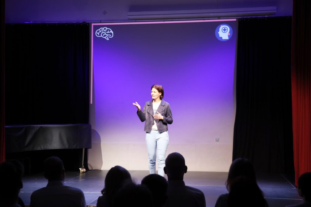 Kirsten Wunderle auf der Bühne vor Publikum, im Hintergrund eine Leinwand mit Gehirn-Symbolen